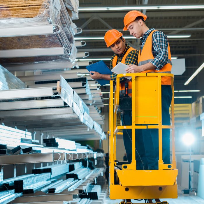 multicultural workers with digital tablet and clipboard standing on scissor lift near shelves with